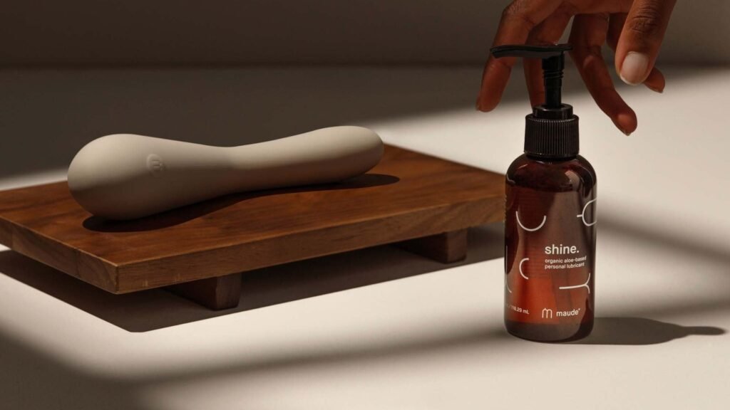 Authentic photograph of the Maude Spot curved vibrator resting on a wooden tray beside Maude Shine organic lubricant in warm natural sunlight
